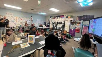 A vibrant classroom filled with students, teachers, and parents engaging in a multilingual writing celebration. A large screen displays student work as attendees participate in a lively learning environment.