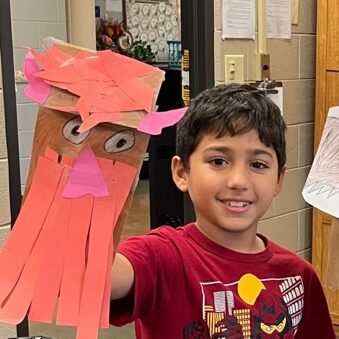 A child holding up a paper puppet or craft creation with a red body and pink details. The child is smiling and wearing a red shirt with a design on it.