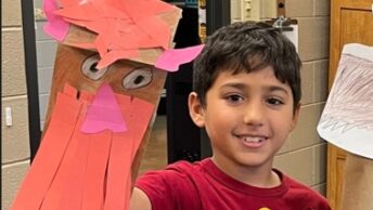 A child holding up a paper puppet or craft creation with a red body and pink details. The child is smiling and wearing a red shirt with a design on it.