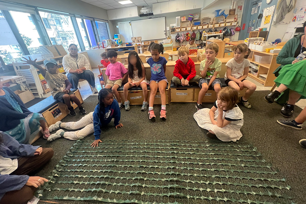 A group of first graders and their teacher sitting around plastic-mesh loom on the floor
