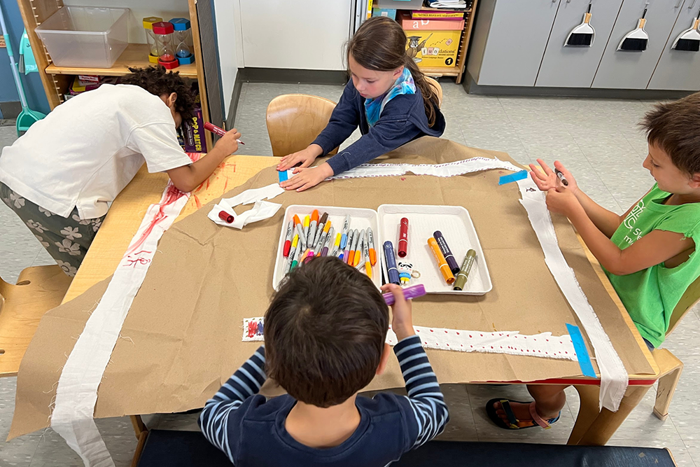 First graders working writing on strips of cloth at a table