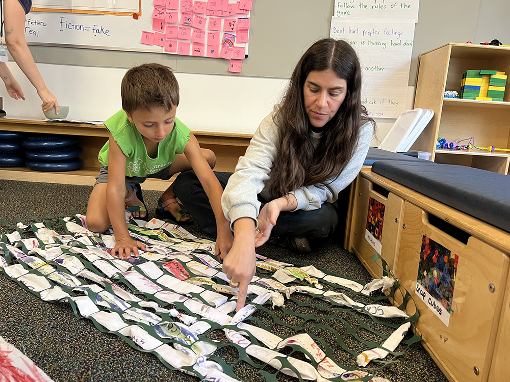 a first-grade student weaves cloth strips in a loom with his art teacher